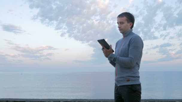 Um homem caminha ao longo da praia perto do oceano com o tablet. Ele verifica mensagens na rede social e navega fotos . — Vídeo de Stock