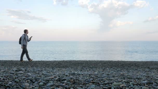 Ein Mann läuft mit dem Tablet am Strand in der Nähe des Meeres entlang. Es ebnet den Weg auf dem Tablet. — Stockvideo