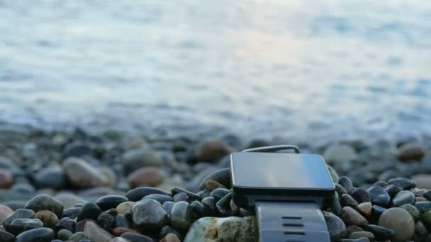 Θολή έξυπνες ρολόγια είναι κοντά στην παραλία με βότσαλο στον ωκεανό. Το ρολόι αντανακλά τα σύννεφα του ουρανού. Γκρο πλαν. — Αρχείο Βίντεο