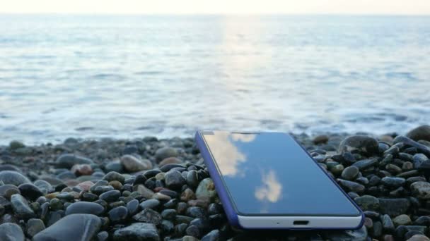 De mobiele telefoon gaat over het kiezelstrand van de Oceaan. Rustige golven tegen de kust slaan. concept van telecommunicatie — Stockvideo