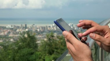 Yüksek katlı binalar ve sıradan evler ile modern bir şehir. Bir adam ön planda telefonunuzda ileti denetler. Yakın çekim. Orman ve deniz arkasında