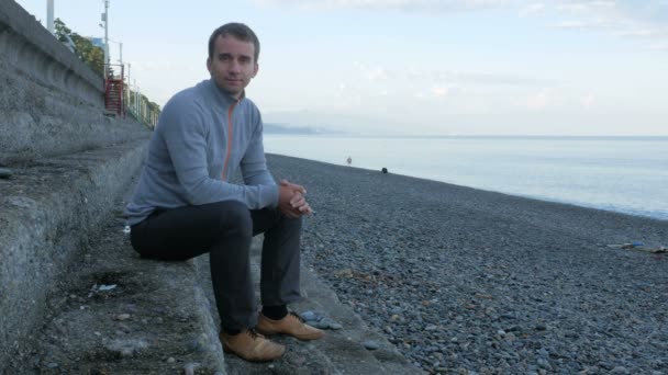 Un homme assis sur les marches près de la plage de galets de l'océan. Il regarde la caméra et sourit — Video