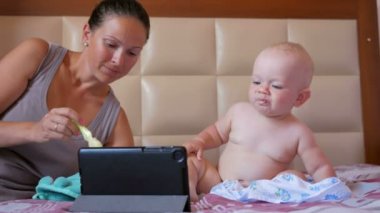 Genç anne şirin bebek kaşığı yulaf lapası ile besleme. Bebek yatak ve bakışları çizgi film Tablet Pc. Tiny üzerinde bir yıl oturma