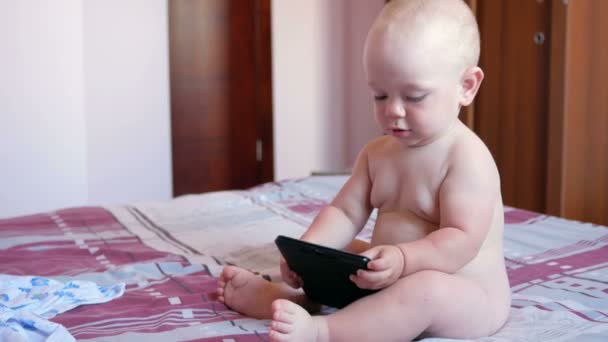 Lindo niño sentado en la cama y mira dibujos animados en la tableta PC. Pequeño bebé de un año sin ropa — Vídeos de Stock