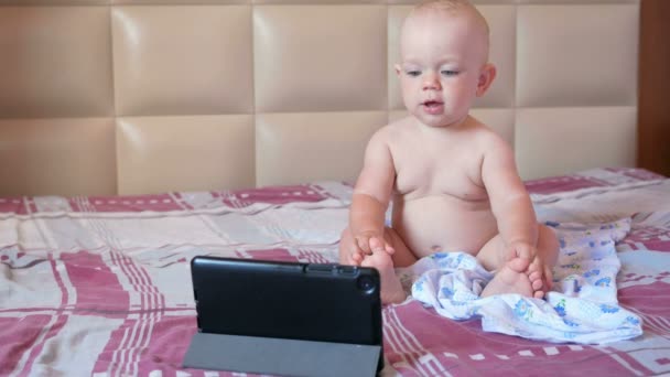 Nettes Kind sitzt auf dem Bett und starrt Cartoons auf den Tablet-PC. Winziges einjähriges Baby ohne Kleidung — Stockvideo