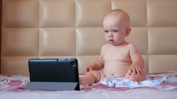 Nettes Kind sitzt auf dem Bett und starrt Cartoons auf den Tablet-PC. Winziges einjähriges Baby ohne Kleidung — Stockvideo