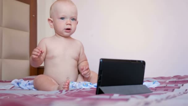 Nettes Kind sitzt auf dem Bett und starrt Cartoons auf den Tablet-PC. Winziges einjähriges Baby ohne Kleidung — Stockvideo