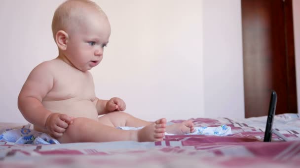 Criança bonito sentado na cama e olha desenhos animados no PC tablet. Pequeno bebê de um ano sem roupas — Vídeo de Stock