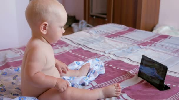 Χαριτωμένο παιδί κάθεται στο κρεβάτι και κοιτάζει επίμονα κινούμενα σχέδια στο tablet Pc. Tiny μωρό ενός έτους χωρίς ρούχα — Αρχείο Βίντεο