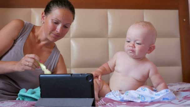 Młoda matka karmienia cute baby z łyżki kaszy. Dziecko siedzi na łóżku i patrzy bajki na tablecie Pc. Tiny jeden rok — Wideo stockowe