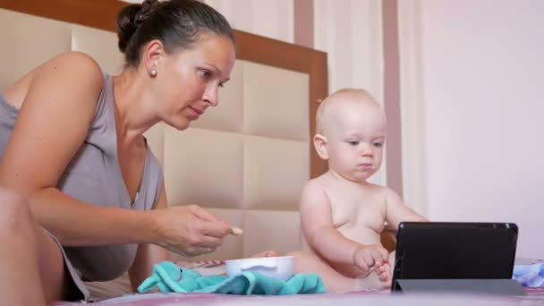 Молодая мама кормит милого ребенка ложкой каши. Ребенок сидит на кровати и смотрит мультики на планшетном компьютере. Крошечный год — стоковое видео