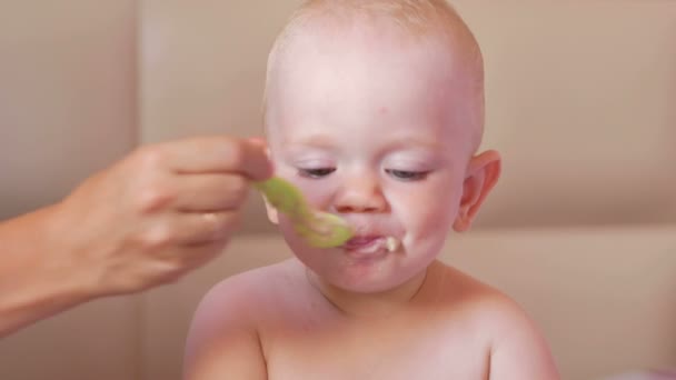 Junge Mutter füttert süßes Baby mit einem Löffel Brei. Baby sitzt auf dem Bett und starrt Karikaturen auf den Tablet-PC. Ein winziges Jahr. Nahaufnahme — Stockvideo