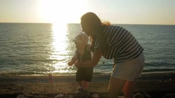 Młoda matka chodzenie z cute dziecko nad brzegiem oceanu o zachodzie słońca. Dziecko nadal jest bardzo słaby, a moja matka przebiegów posiada. Pojęcie pierwsze kroki w życiu — Wideo stockowe