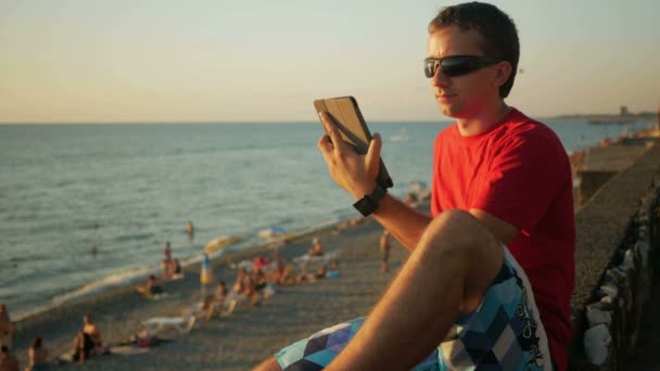 Joven hombre guapo en gafas de sol relajante cerca de la playa del mar con tableta de ordenador. Chequea mensajes en las redes sociales al atardecer . — Vídeo de stock