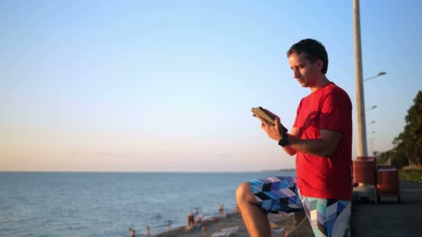 Όμορφος νεαρός χαλάρωσης κοντά στην παραλία της θάλασσας με τον υπολογιστή tablet. Αυτός ελέγχει μηνυμάτων για τα κοινωνικά δίκτυα στο ηλιοβασίλεμα. — Αρχείο Βίντεο