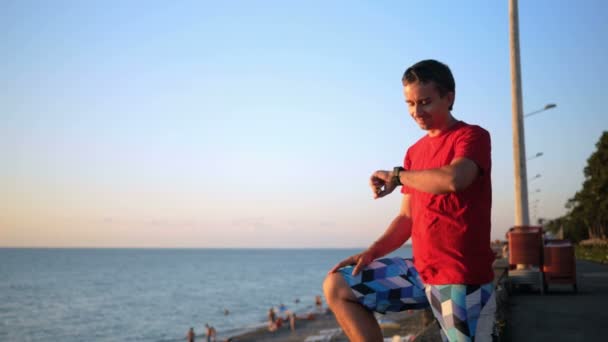 Jovem homem bonito relaxando perto da praia do mar com relógio inteligente. Ele verifica mensagens nas redes sociais ao pôr do sol no smartwatch . — Vídeo de Stock