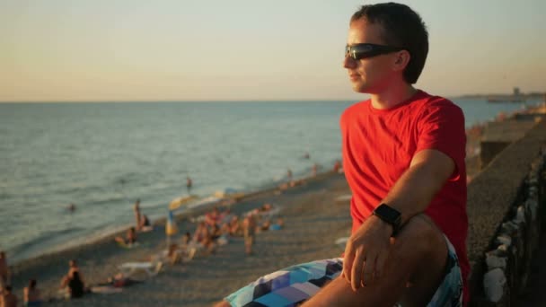 Όμορφος νεαρός με γυαλιά ηλίου χαλάρωσης κοντά στην παραλία στη θάλασσα στο ηλιοβασίλεμα. Θαυμάζει το ηλιοβασίλεμα και το νερό. Μετά γυρίζοντας το κεφάλι του και εξετάζοντας τη φωτογραφική μηχανή — Αρχείο Βίντεο