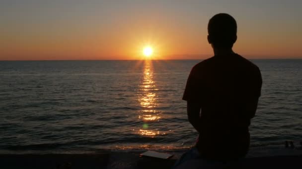 Silhueta de um homem ao pôr-do-sol na praia do oceano. Um homem admira que o sol alaranjado esteja quase posto . — Vídeo de Stock