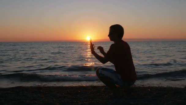 Silueta hombre con tableta digital en las manos al atardecer playa. El sol casi se pone detrás del océano . — Vídeos de Stock