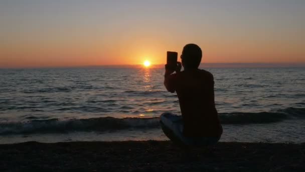 Silueta hombre con tableta digital tomando fotos al atardecer playa. El sol casi se pone detrás del océano . — Vídeos de Stock