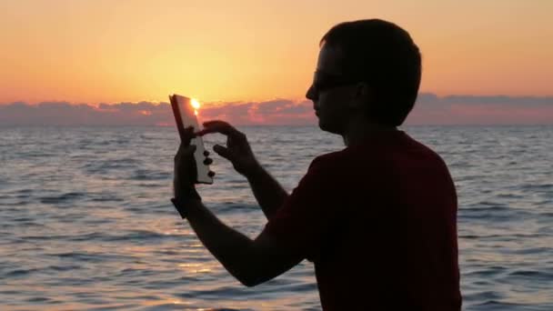 Silhouet man met digitale tablet in handen op sunset beach. De zon ligt bijna achter de Oceaan. — Stockvideo