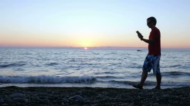 Σιλουέτα ενός ανθρώπου στο ηλιοβασίλεμα περνά κατά μήκος της προκυμαίας με έναν υπολογιστή tablet. Αυτός σταματά, ελέγξτε το μήνυμα και να κινείται. έννοια των διακοπών — Αρχείο Βίντεο