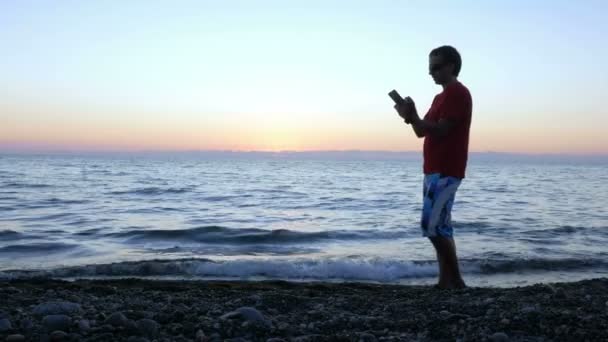 La silhouette d'un homme au coucher du soleil passe le long du front de mer avec une tablette. Il s'arrête, vérifie le message et passe à autre chose. concept de vacances — Video