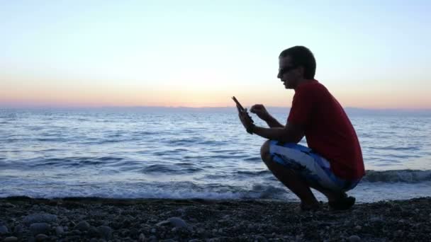 Sylwetka człowieka o zachód słońca siedząc na nabrzeżu przy użyciu komputera typu tablet. Sprawdza wiadomości z jego dziewczyna na portalach społecznościowych. koncepcja wakacje — Wideo stockowe