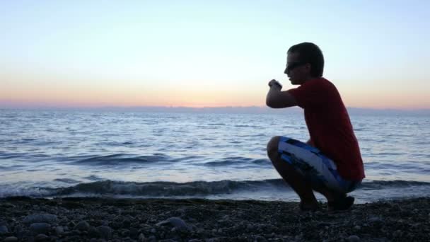 Sylwetka człowieka o zachód słońca siedząc na nabrzeżu i oferujący smartwatch. Sprawdza wiadomości ze swoją dziewczyną na portalach społecznościowych na inteligentny zegarek. koncepcja wakacje — Wideo stockowe