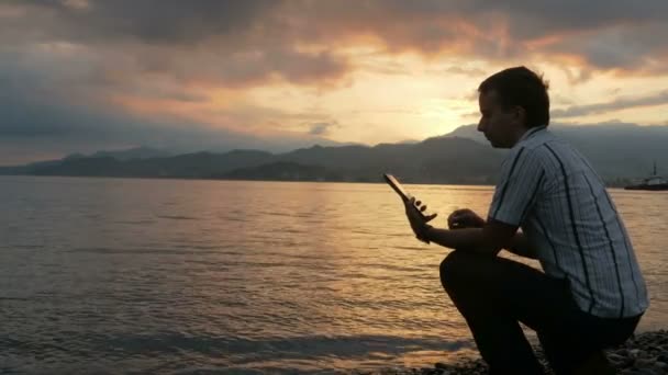 Un homme en chemise vérifie les messages sur la tablette pendant le lever du soleil sur la plage de l'océan. Merveilleuses couleurs du ciel et le lever du soleil de derrière les montagnes . — Video