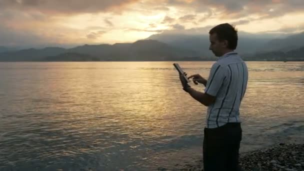 Um homem de camisa verifica as mensagens no tablet durante o nascer do sol na praia do oceano. Maravilhosas cores do céu e do nascer do sol por trás das montanhas . — Vídeo de Stock