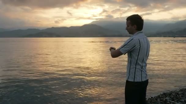 Um homem de camisa verifica as mensagens no relógio inteligente durante o nascer do sol na praia do oceano e das montanhas. Há um ditado mensagens de voz. Cores deslumbrantes do céu e do sol nascente — Vídeo de Stock