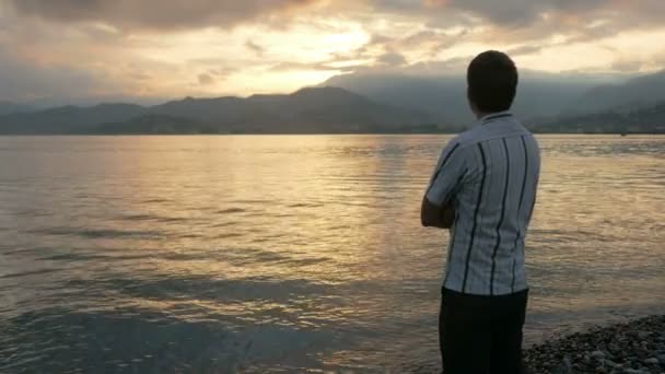 L'uomo guarda con una camicia che guarda l'alba sulla spiaggia dell'oceano. Il sole sorge da dietro le montagne. Il concetto di ammirazione della natura — Video Stock