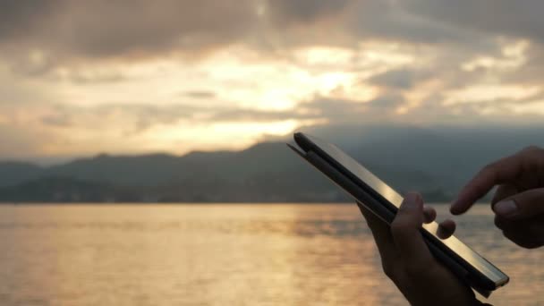 Un hombre comprueba los mensajes en la tableta durante el amanecer en la playa del océano. Impresionantes colores del cielo y el sol naciente. Primer plano — Vídeos de Stock