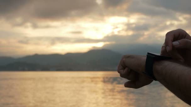 Un uomo controlla i messaggi sull'orologio intelligente durante l'alba sulla spiaggia dell'oceano. Meravigliosi colori del cielo e il sorgere del sole da dietro le montagne. Primo piano — Video Stock