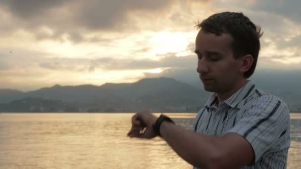 Un homme en chemise vérifie les messages sur la montre intelligente pendant le lever du soleil sur la plage de l'océan et les montagnes. Il y a un dictée messages vocaux. Superbes couleurs du ciel et du soleil levant — Video