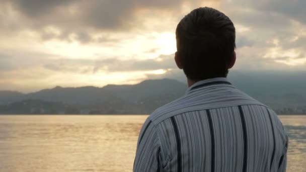 De man kijkt in een shirt dat kijken naar de zonsopgang op het strand van de Oceaan. De stijgingen van de zon achter de bergen. Het concept van natuur bewondering — Stockvideo