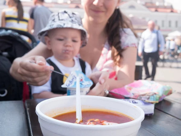 Прекрасний малюк ходить з матір'ю на міське свято. Хлопчик їсть суп зі сметаною на вулиці. Дитяча стильно одягнена метелик і шапка Ліцензійні Стокові Зображення