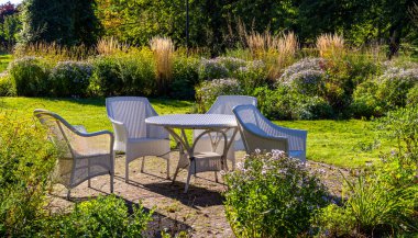 Resim, Britzer Garden 'da dört beyaz sandalyesi ve yuvarlak bir masası olan iyi korunmuş bir oturma alanını gösteriyor. Çevre sonbahar renkli çimen ve çalılarla karakterize edilir; arka planda üç büyük taş heykel ve ağaçlar vardır.