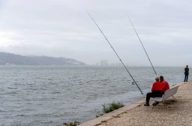 Ponte 25 de Abril 'deki balıkçılar, Portekiz' deki 3,2 km uzunluğunda bir köprü kompleksi, Tagus Nehri üzerindeki 2278 metre uzunluğunda asma köprüsü, bulutlu ve yağmurlu hava, birleşik yol ve demiryolu trafiğine sahip üçüncü en uzun asma köprü, Lizbon