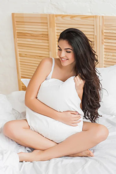 Mujer sonriente sosteniendo una almohada mientras está sentada en su cama — Foto de Stock