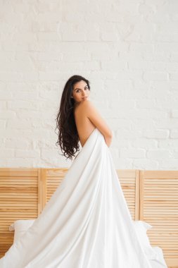 Beyaz yatak halfturned üzerinde duran, kameraya bakarak seksi kadın