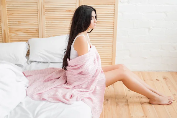 Schöne junge Brünette mit rosa Decke bedeckt auf dem Bett sitzend — Stockfoto