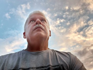 Yaşlı adam gözlerinde umutla yukarı bakıyor. Arkaplanda bulutlu bir gökyüzü.