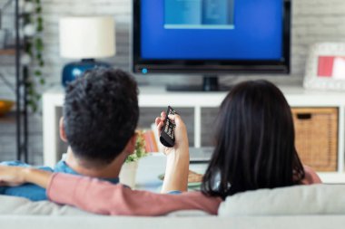 Evdeki kanepede televizyon izlerken uzaktan kumandayla kanalı değiştiren rahat genç çiftin çekimi..