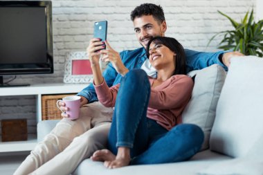 Mutlu genç bir çiftin evdeki koltukta otururken akıllı telefonuyla video konuşması..