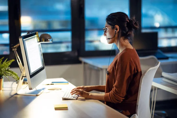 オフィスに座っているイヤホンと話をしながら コンピュータを操作する美しい若いビジネス女性のショット — ストック写真