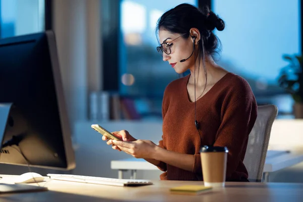 オフィスに座ってコンピュータと仕事をしながら携帯電話でメッセージを送信する美しい若いビジネス女性のショット — ストック写真