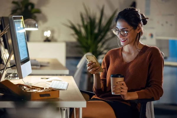 オフィスに座ってコンピュータと仕事をしながら携帯電話でビデオ通話を持っている美しい若いビジネス女性のショット — ストック写真