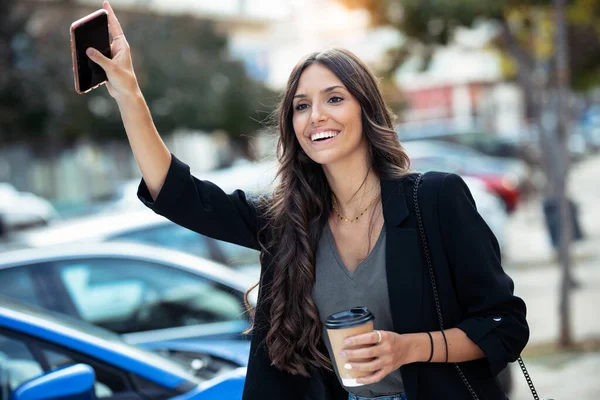 スマートフォンとカップコーヒーを持ちながら手を振って笑顔の若いビジネス女性のショットが通りを歩いて — ストック写真